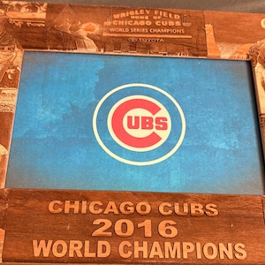 Puede incluir: Marco de madera con el logotipo de los Chicago Cubs y escenas de béisbol. El marco incluye el texto "Wrigley Field Home of Chicago Cubs World Series Champions" y "Chicago Cubs 2016 World Champions."