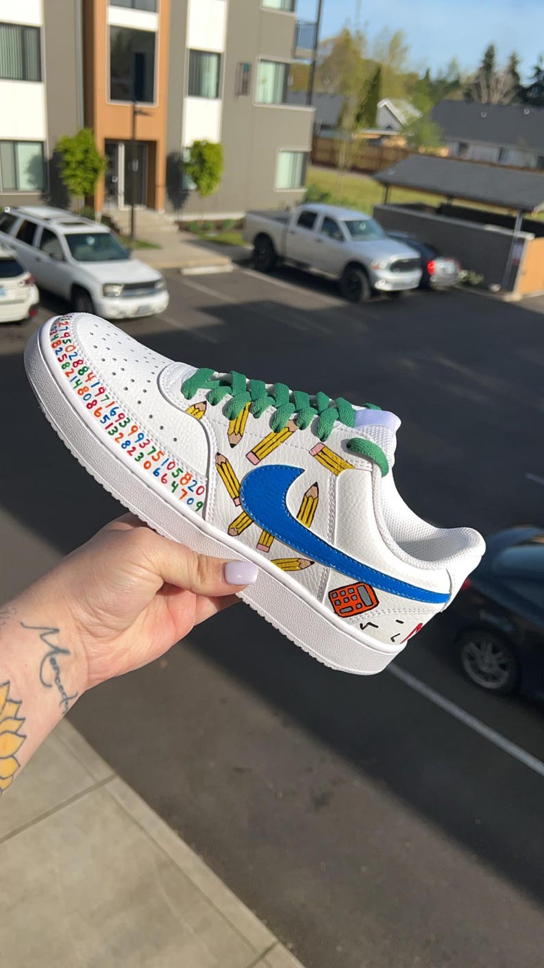 May include: White sneaker with blue Nike swoosh, green laces, and pencil and calculator graphics. The shoe features a colorful number sequence along the side. A fun, school-themed design.