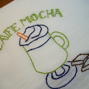 May include: White embroidered cloth with the words "CAFE MOCHA" in green thread. The design features a coffee cup with whipped cream, a straw, and coffee beans, all embroidered in blue, green, orange, and brown thread.