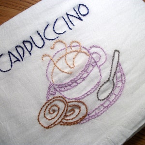 May include: White embroidered kitchen towel with the word "CAPPUCCINO" in dark blue thread. The towel features an embroidered cappuccino cup, spoon, and two cookies in shades of purple, tan, and brown.