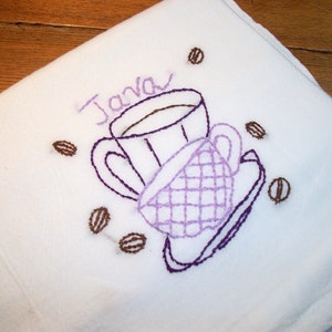 May include: White embroidered towel featuring a coffee cup and saucer design with the word "Java" in purple thread. Surrounding the cups are embroidered coffee beans in brown. The towel is folded, showing a corner.