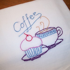 May include: White embroidered kitchen towel featuring a coffee cup, cupcake, and the word "Coffee" in blue thread. The coffee cup has a blue and white checkered design. The cupcake has a cherry on top.