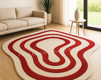 May include: A cream-colored rug with a unique, organic shape and bold red wavy stripes. The rug is placed on a hardwood floor in a living room setting, with a sofa and side table visible in the background. The text "FAIZAHANDMADERUGS" is printed on the rug.