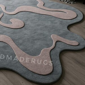 Pu&ograve; includere: Un tappeto moderno con un design astratto. Il tappeto presenta un colore di base grigio-blu con forme organiche fluide in rosa tenue. Il testo "DMADERUGS" &egrave; visibile sulla superficie del tappeto. Il tappeto &egrave; posato su un pavimento in legno.