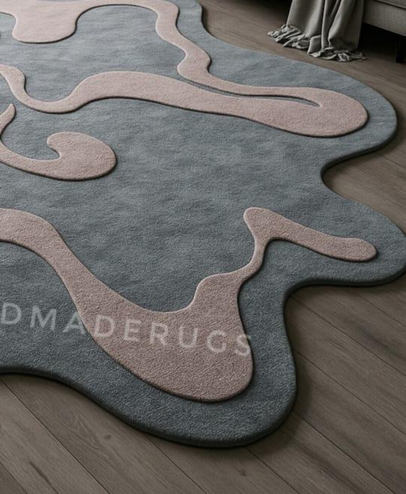 Pu&ograve; includere: Un tappeto moderno con un design astratto. Il tappeto presenta un colore di base grigio-blu con forme organiche fluide in rosa tenue. Il testo "DMADERUGS" &egrave; visibile sulla superficie del tappeto. Il tappeto &egrave; posato su un pavimento in legno.