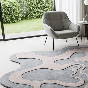 Pu&ograve; includere: Un tappeto moderno con un design astratto nei toni del grigio e del rosa chiaro. Il tappeto &egrave; posizionato su un pavimento in legno chiaro, con una poltrona grigia e una finestra sullo sfondo. La forma unica del tappeto aggiunge un tocco contemporaneo alla stanza.