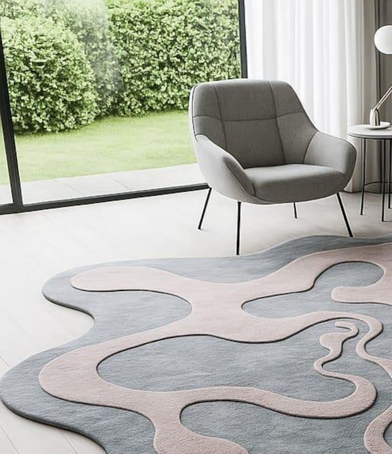 Pu&ograve; includere: Un tappeto moderno con un design astratto nei toni del grigio e del rosa chiaro. Il tappeto &egrave; posizionato su un pavimento in legno chiaro, con una poltrona grigia e una finestra sullo sfondo. La forma unica del tappeto aggiunge un tocco contemporaneo alla stanza.