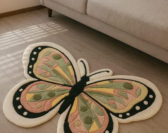 Puede incluir: Una alfombra con forma de mariposa con un borde color crema y un interior colorido. Las alas de la alfombra presentan un patrón de secciones rosas, verdes y amarillas, con detalles en negro y puntos blancos. La alfombra está colocada sobre un suelo de madera.