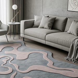 Pu&ograve; includere: Un moderno soggiorno con un divano grigio, cuscini e un tappeto unico. Il tappeto ha una base grigio-azzurra con forme rosa astratte. Un tavolino con lampada e opere d'arte completano la scena. Tonalit&agrave; di grigio.