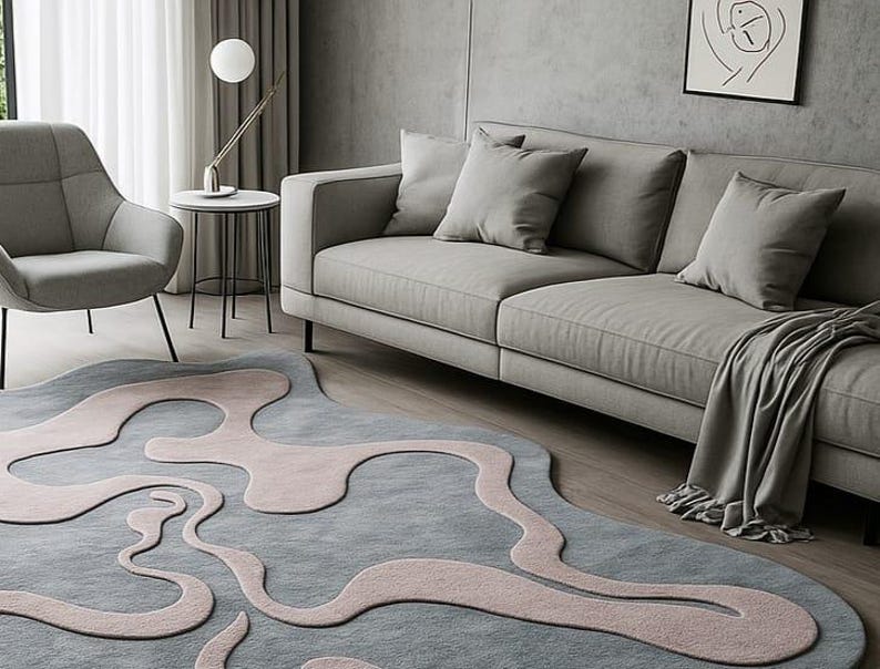 Pu&ograve; includere: Un moderno soggiorno con un divano grigio, cuscini e un tappeto unico. Il tappeto ha una base grigio-azzurra con forme rosa astratte. Un tavolino con lampada e opere d'arte completano la scena. Tonalit&agrave; di grigio.