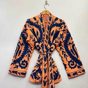 Puede incluir: Una chaqueta de manga larga en un tono naranja brillante con un patrón floral azul marino oscuro en toda la prenda. Incluye un cinturón a juego en la cintura, un diseño frontal abierto y mangas anchas, todo exhibido en una percha de madera.