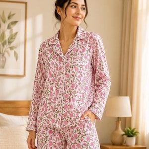 May include: A long-sleeved button-down pajama top and matching pants set featuring a white base with a repeating pink floral pattern and green stems. The top includes a notched collar, a single chest pocket, and white piping along the edges.