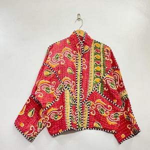 Può includere: Giacca trapuntata rossa con collo alto, decorata con disegni paisley e floreali gialli e verdi. La giacca ha finiture a righe bianche e nere. Appesa a una gruccia di legno, presenta un design colorato e originale.