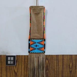 Puede incluir: Una bolsa de gamuza marrón con un diseño de cuentas y flecos. La sección de cuentas presenta un patrón geométrico azul y naranja con una cruz en el centro. Los lados de la bolsa tienen detalles de cuentas rojas, amarillas y naranjas.