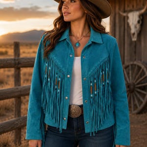 May include: A bright turquoise suede jacket featuring long fringe detailing across the chest and sleeves, accented with small wooden and blue beads. The jacket has a classic collar, silver-toned metal snap buttons, and is worn over a white top.