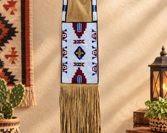 May include: A tan suede wall hanging featuring a rectangular beaded panel with white, maroon, and blue geometric patterns, including a central cross motif. The top is bordered by a beaded strip, and the bottom consists of long, thin suede fringe.
