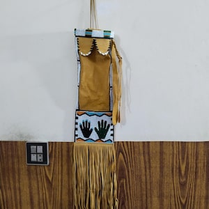 May include: A handmade, beaded pouch with a tan leather body and fringe. The top features a beaded design in white, blue, and green. The bottom section displays two hand prints. The pouch is suspended by a cord.