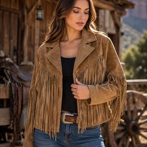 May include: A tan suede jacket featuring long fringe detailing across the chest, back, and sleeves. The jacket has a notched lapel collar, a button-front closure, and long sleeves. It is worn over a black ribbed top and blue denim jeans.