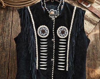 May include: A black suede vest featuring long fringe along the armholes, white beaded accents, and circular beaded medallions on the chest. The vest has a high collar with colorful trim, a front button closure, and white braided leather piping.