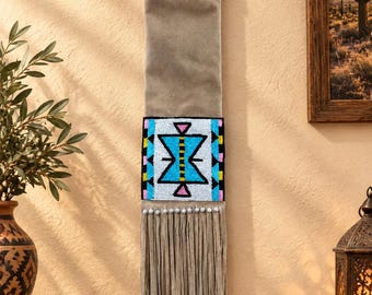 May include: A vertical wall hanging made of tan suede-like material featuring a top band of white, black, and blue seed beads. A central rectangular panel displays a geometric pattern in bright blue, white, pink, and black beads, finished with long fringe.