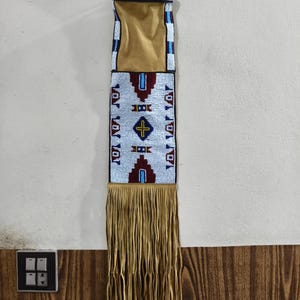 Pode incluir: Uma bolsa de contas com franjas de couro castanho. A bolsa apresenta um bolso de camurça castanho e é decorada com contas brancas, vermelhas, azuis e amarelas num padrão geométrico. As franjas são de cor castanho claro.