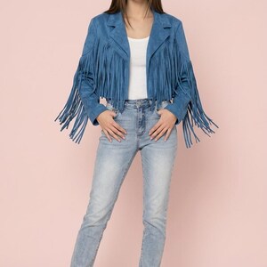 Puede incluir: Chaqueta de ante azul con flecos y corte cropped. La chaqueta presenta largos flecos en las mangas y el dobladillo. La modelo lleva vaqueros de lavado claro y botines color canela. El fondo es de color rosa liso.