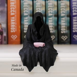 May include: A black figurine depicting a cloaked figure holding a small pink teacup. The figurine is positioned in front of a row of books with the author's name "Rebecca Yarros" on the spines. The text "Made in Canada" is also visible.