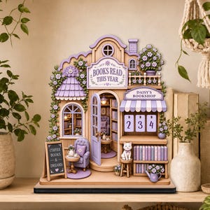 Puede incluir: Una maqueta de librería de madera en miniatura con una combinación de colores morado y blanco. El modelo presenta letreros que dicen "Books Read This Year" y "Daisy's Bookshop". Una pequeña pizarra muestra "Coffee Books & Dreams".