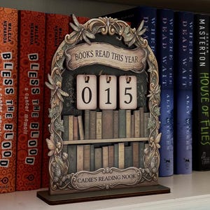 May include: A decorative wooden book tracker featuring an arched frame with dragonfly and fairy motifs. It displays the text "BOOKS READ THIS YEAR" and "CADIE'S READING NOOK" with three hanging number tiles showing "015" above a shelf of miniature books.