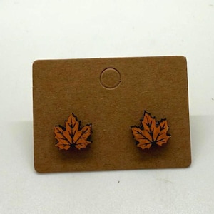 Puede incluir: Un par de pendientes de botón con forma de hoja de arce. Los pendientes son de color naranja con detalles negros, montados en una tarjeta de exhibición marrón. Un accesorio con temática otoñal.