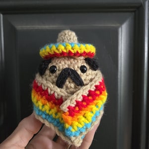 May include: A crocheted pug wearing a colorful sombrero and poncho. The pug's body is beige, with black eyes and nose. The sombrero is yellow, blue, and red. The poncho features stripes of red, orange, yellow, blue, and beige.