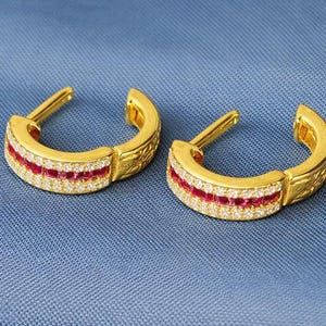 May include: A pair of gold hoop earrings featuring a red and white gemstone design. The earrings are displayed on a blue background.