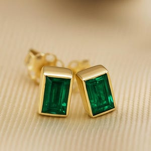 May include: A pair of gold stud earrings with emerald-cut green gemstones. The earrings have a rectangular shape with rounded corners and are set in a gold bezel setting. The background is a soft, neutral color.