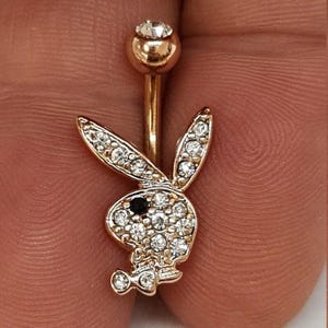 Pode incluir: Anel de umbigo dourado com um design de logotipo de coelho. O coelho é incrustado com cristais transparentes e um olho de cristal preto. A bola superior é dourada com um único cristal transparente.