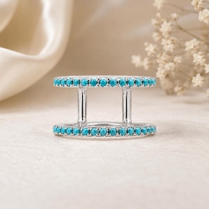 May include: A silver-toned double band ring featuring two parallel rows of small, round, bright turquoise stones. The two bands are connected by two vertical silver-toned bars, creating an open, architectural design with a polished finish.