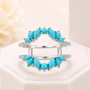 May include: A silver ring with a double band design. The top and bottom bands feature a row of small, clear stones and a series of turquoise-colored, oval-shaped stones. The ring is set against a white background.