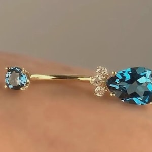 May include: A gold belly button ring with a round blue gemstone on one end and a teardrop-shaped blue gemstone on the other. Small white gemstones are clustered near the teardrop gem.