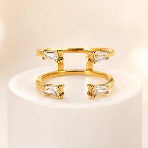 May include: A gold-coloured ring with a distinctive open design. The ring has four rectangular, clear gemstones. It is displayed on a white surface against a soft, neutral background. The ring's design is modern and elegant.