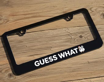 Puede incluir: Marco de matrícula negro con el texto blanco « GUESS WHAT » y un dibujo de un pollo. El marco rectangular está sobre una superficie de madera.