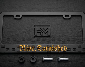 Puede incluir: Un marco de matrícula negro rectangular con acabado mate y un patrón cuadrado repetitivo en el borde inferior. El marco muestra el texto "Rise, Tarnished" en una fuente gótica naranja e incluye dos tapas negras y dos tornillos plateados.