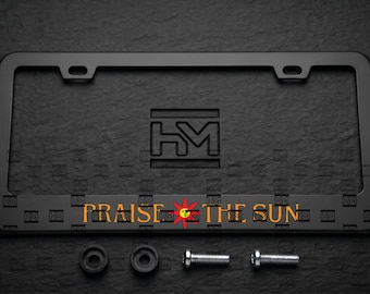 Puede incluir: Un marco de matrícula rectangular de color negro mate con las letras HM en relieve en la parte superior central y el texto PRAISE THE SUN en naranja con un icono de sol rojo en la parte inferior. Incluye dos espaciadores negros y dos tornillos.