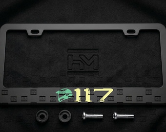 Puede incluir: Un marco de matrícula rectangular negro mate con las letras HM en relieve en la parte superior central. El borde inferior muestra un patrón repetido de pequeños logotipos HM, un icono de casco verde claro y el texto amarillo 117. Incluye dos tornillos.