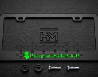 Puede incluir: Marco de matrícula negro con el texto "[Everyone disliked that!]". El marco tiene forma rectangular con esquinas redondeadas y orificios de montaje. Incluye accesorios de montaje. El fondo es una superficie oscura y texturizada.