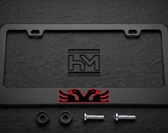 Puede incluir: Un marco de matrícula rectangular negro mate con un logotipo HM grabado en el centro superior y un gráfico de águila bicéfala negra con un fino contorno rojo en la parte inferior. Incluye dos espaciadores de goma negros y dos tornillos metálicos.