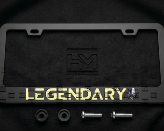 Puede incluir: Un marco de matrícula rectangular negro con la palabra LEGENDARY en letras mayúsculas amarillas, acompañada de un pequeño icono de calavera. El marco incluye dos orificios de montaje en la parte superior, dos tapas de tornillos negras y dos tornillos.