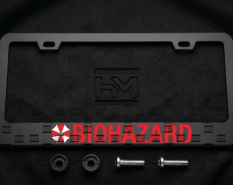 Puede incluir: Un marco de matrícula rectangular negro mate con el logotipo de Biohazard en forma de paraguas rojo y blanco y la palabra BIOHAZARD en letras mayúsculas rojas. El marco incluye dos espaciadores circulares negros y dos tornillos metálicos plateados.