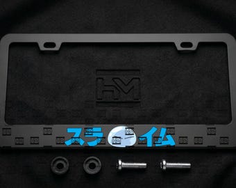 Puede incluir: Un marco rectangular negro para matrícula con las letras HM en relieve en el centro y un patrón repetitivo de pequeños logotipos HM a lo largo del borde inferior. El marco incluye dos espaciadores circulares negros y dos tornillos de montaje de metal plateado.
