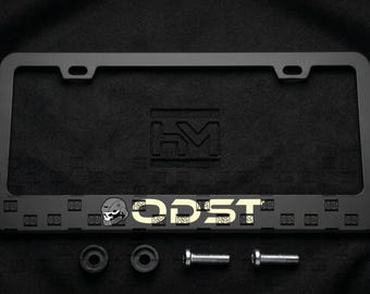 Puede incluir: Un marco de matrícula rectangular negro mate con un patrón cuadrado repetitivo en el borde inferior. El texto ODST está impreso en color beige claro junto a un icono de casco con forma de calavera. Dos espaciadores negros y dos tornillos metálicos.