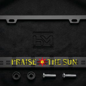 May include: A matte black rectangular number plate frame featuring the yellow text "PRAISE THE SUN" with a red sun icon in the centre. The frame includes two mounting holes at the top, two black circular grommets, and two silver metal screws.