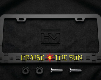 Puede incluir: Un marco de matrícula rectangular negro mate con el texto amarillo "PRAISE THE SUN" y un icono de sol rojo en el centro. El marco incluye dos orificios de montaje en la parte superior, dos arandelas circulares negras y dos tornillos metálicos plateados.